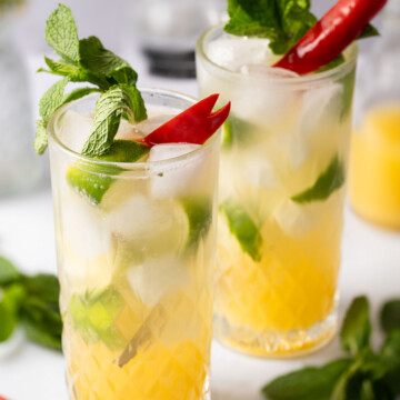 Two glasses of mango punch with mint, lime and red chilli garnishes.