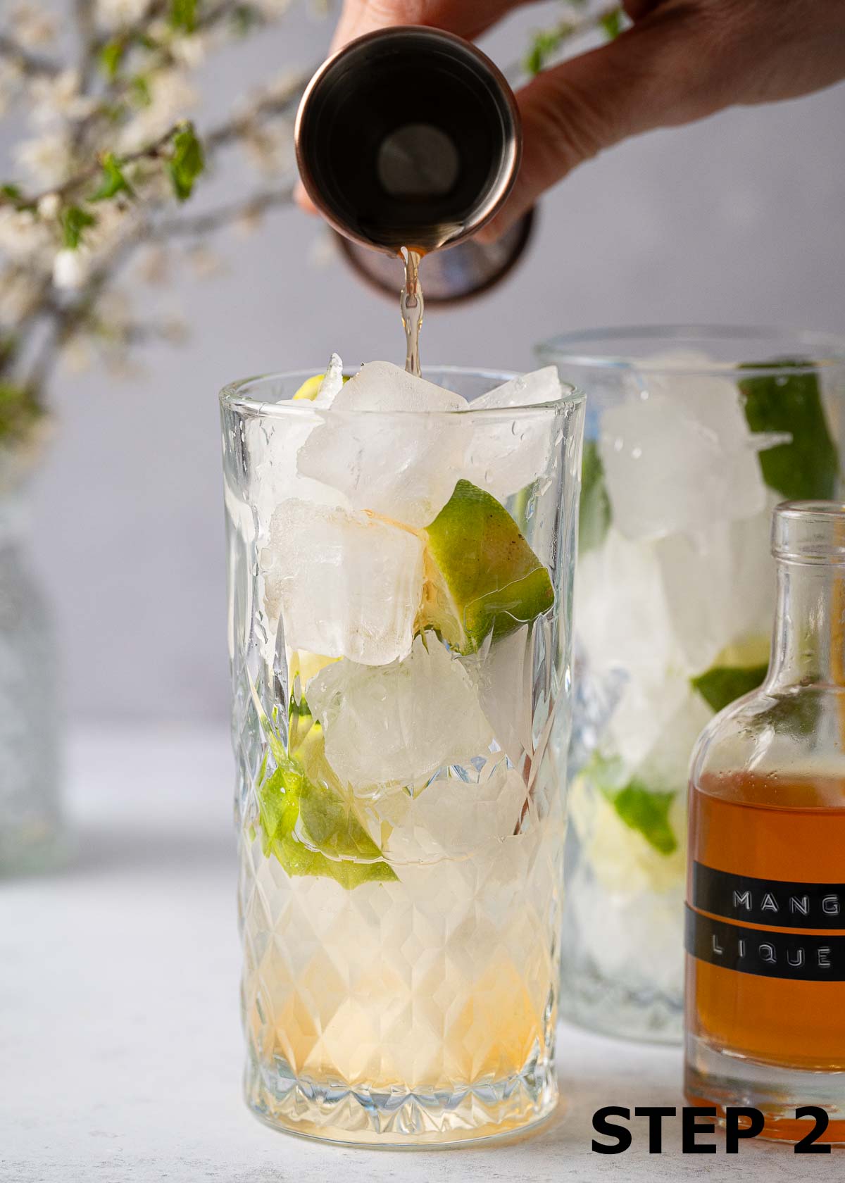 A person pouring mango liqueur into a highball glass with ice and lime in it.