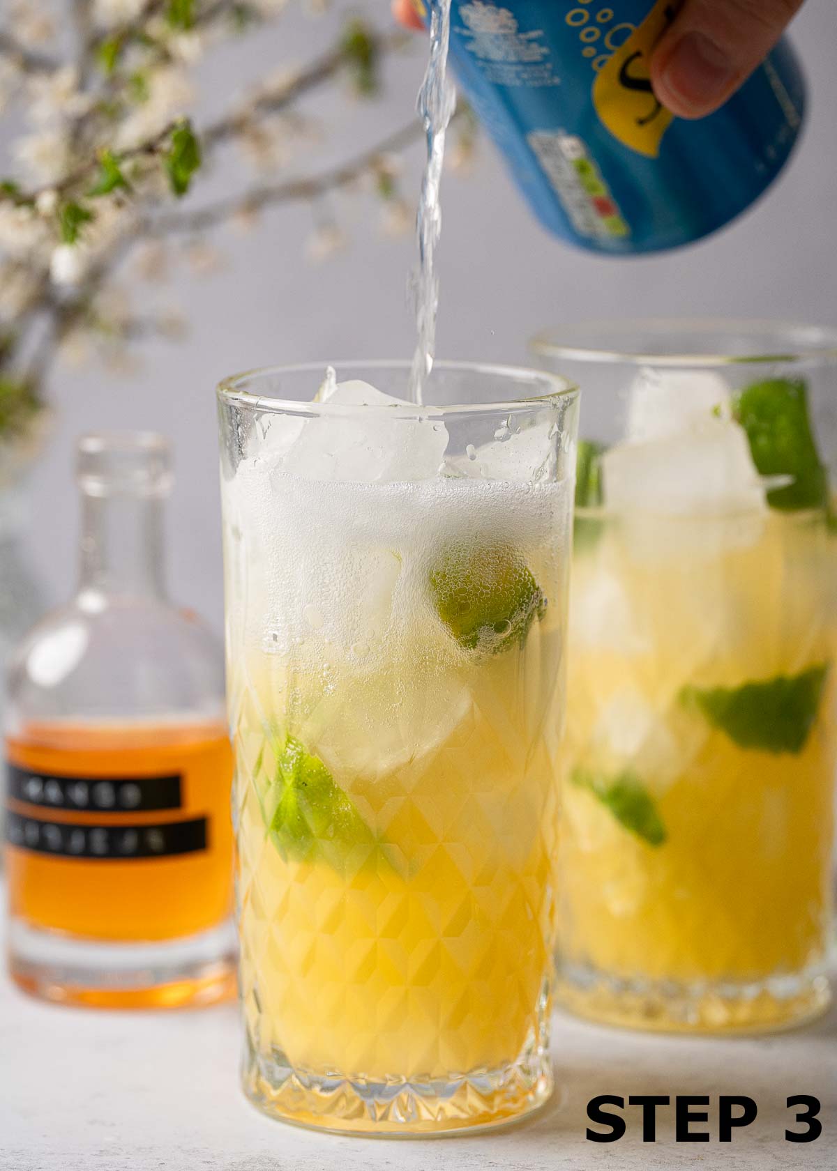 A person pouring lemonade from a can into a mango punch cocktail in a glass.