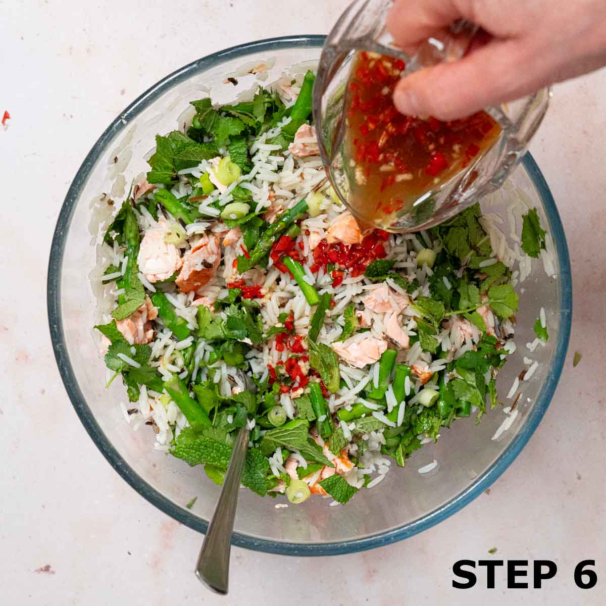 A person pouring spicy Thai-style dressing over rice, flaked salmon, asparagus and herbs.