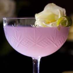 A violet cocktail in a vintage coupe glass garnished with a curl of lime and a white flower.