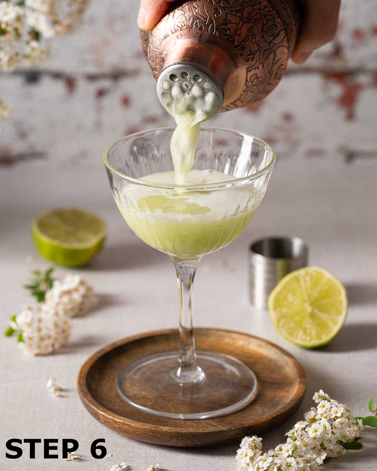 A person pouring a green basil gin cocktail into a coupe glass.