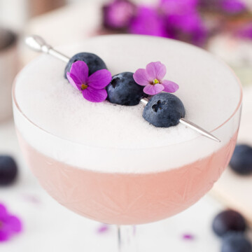 A blueberry gin cocktail with egg white foam garnished with blueberries & flowers.