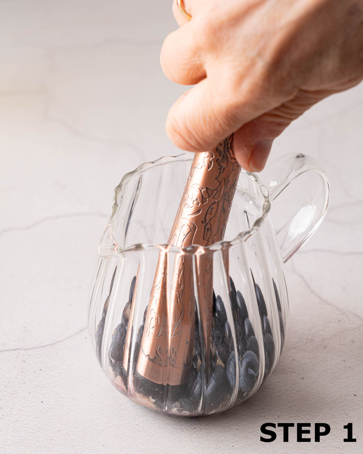 A person muddling fresh blueberries in a glass jug with a cocktail muddler.