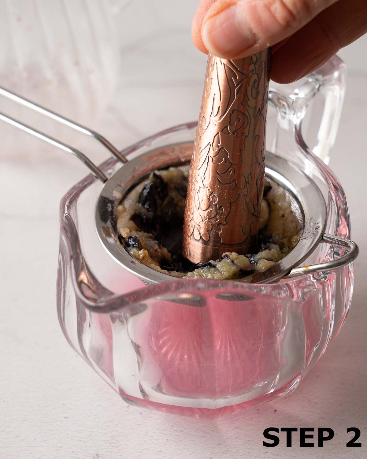 A person pressing blueberries and gin through a sieve into a jug using a cocktail muddler.