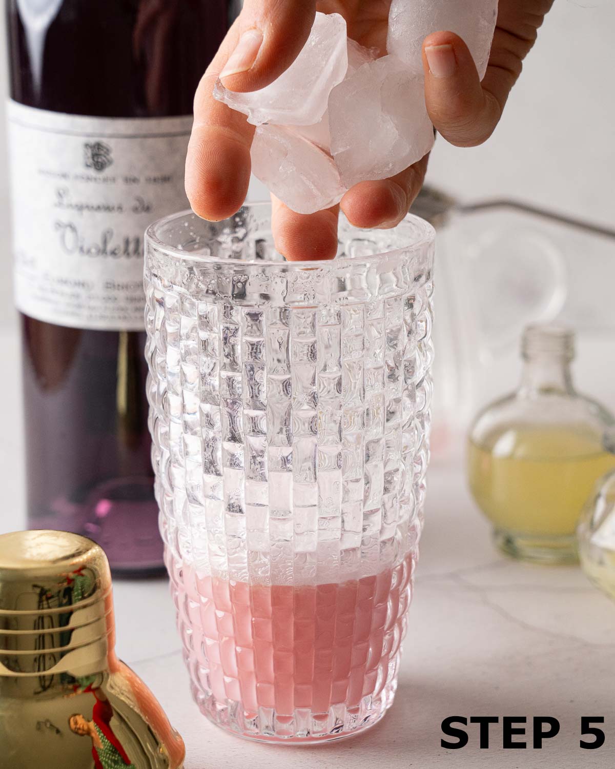 A person adding ice to a shaker holding a blueberry creme de violette cocktail.