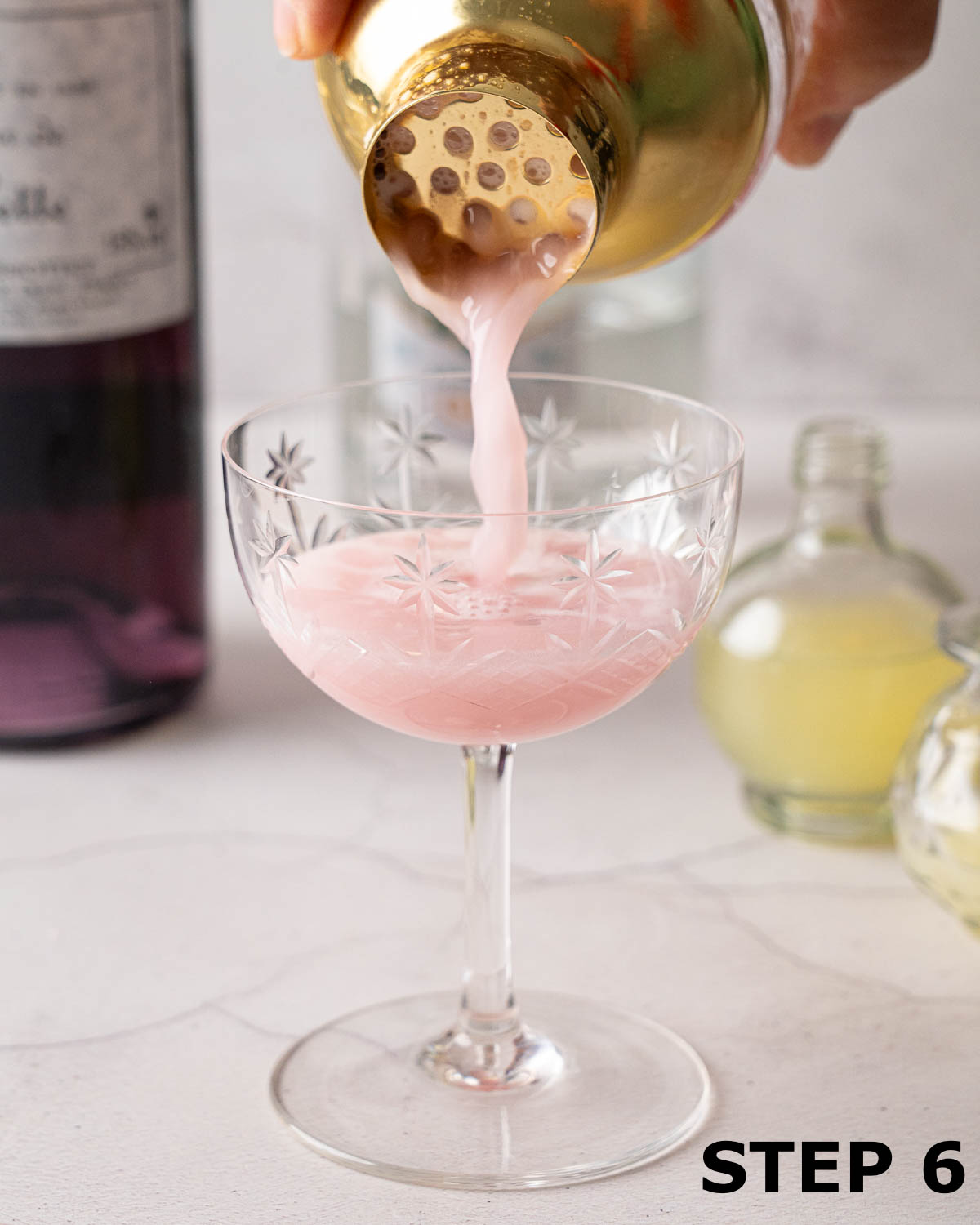 A person straining a blueberry gin cocktail into a vintage coupe glass.