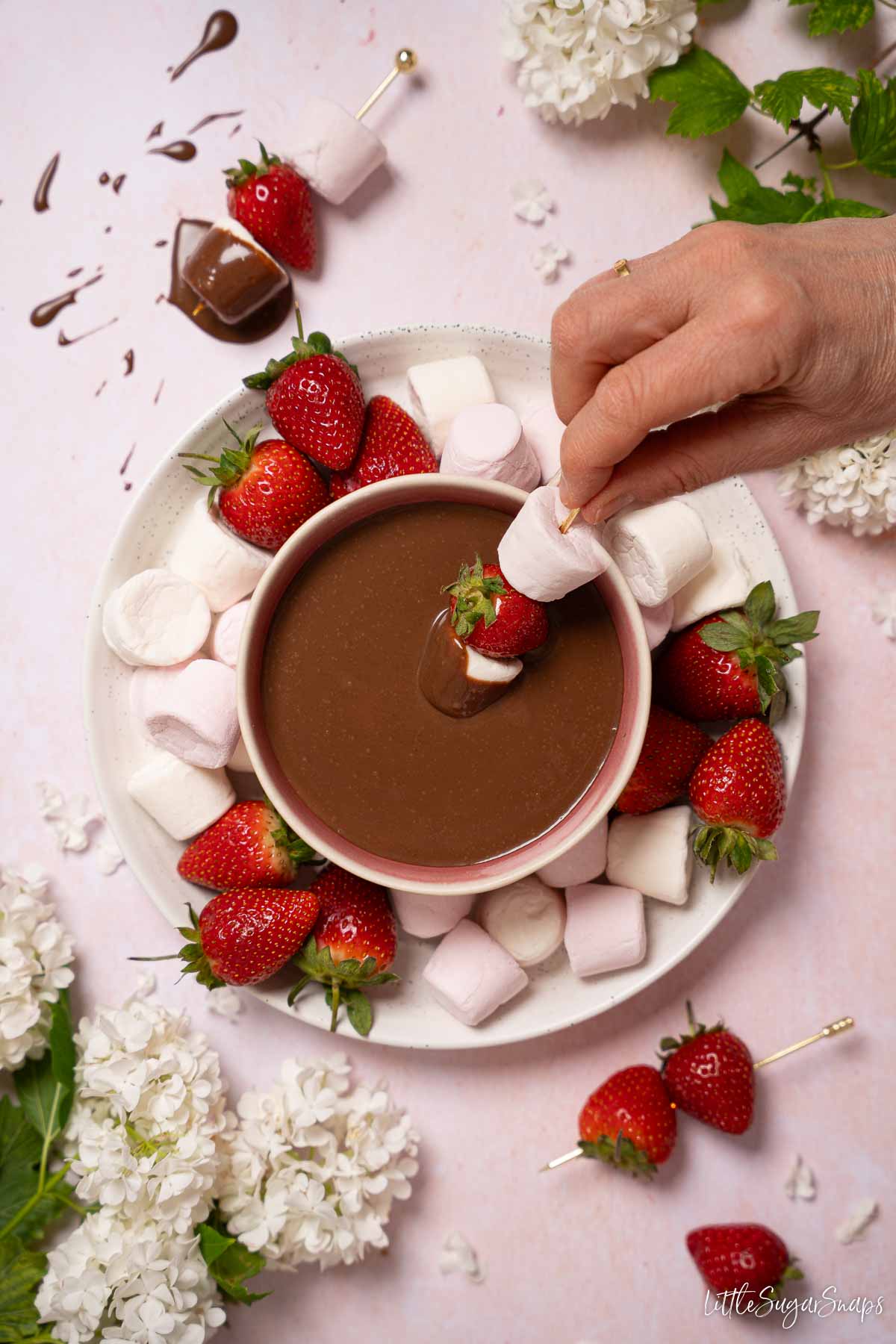 Plated marshmallows and strawberries with chocolate dipping sauce on a plate and someone dipping skewered marshmallows into it.