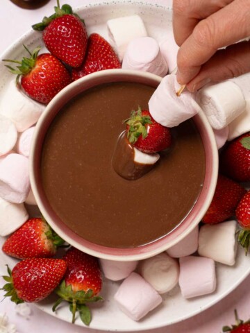 A person dipping skewered marshmallows and strawberries into chocolate dip.