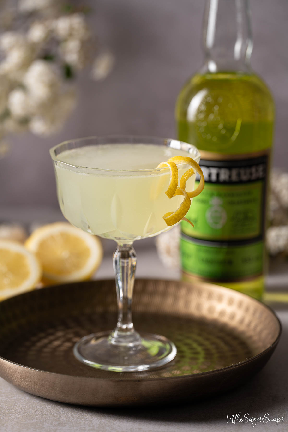 A gin and Green Chartreuse cocktail garnished with lemon zest spirals with a liqueur bottle behind.
