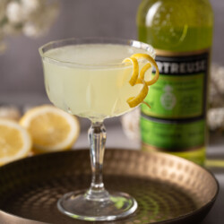 A Green Chartreuse cocktail garnished with lemon zest spirals with a bottle of liqueur behind.