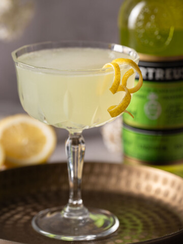 A Green Chartreuse cocktail garnished with lemon zest spirals with a bottle of liqueur behind.