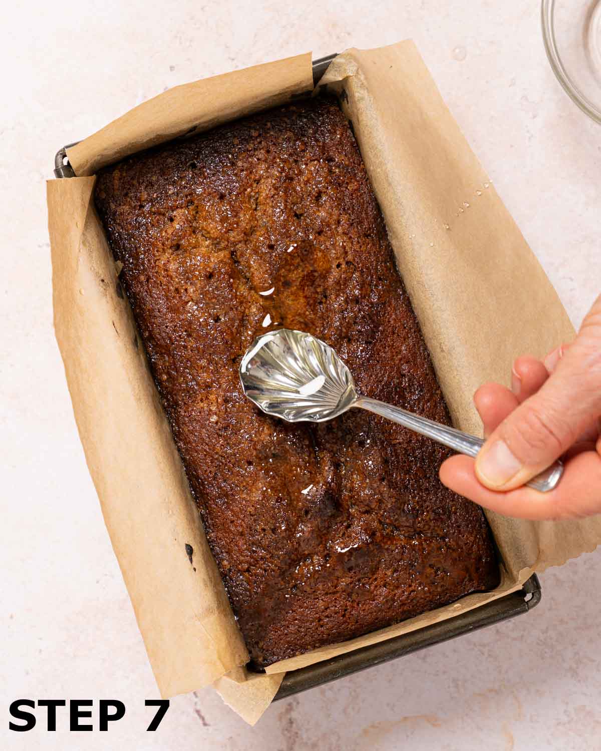 A person spooning stem ginger syrup over the top of a ginger and date cake still in the baking tin.