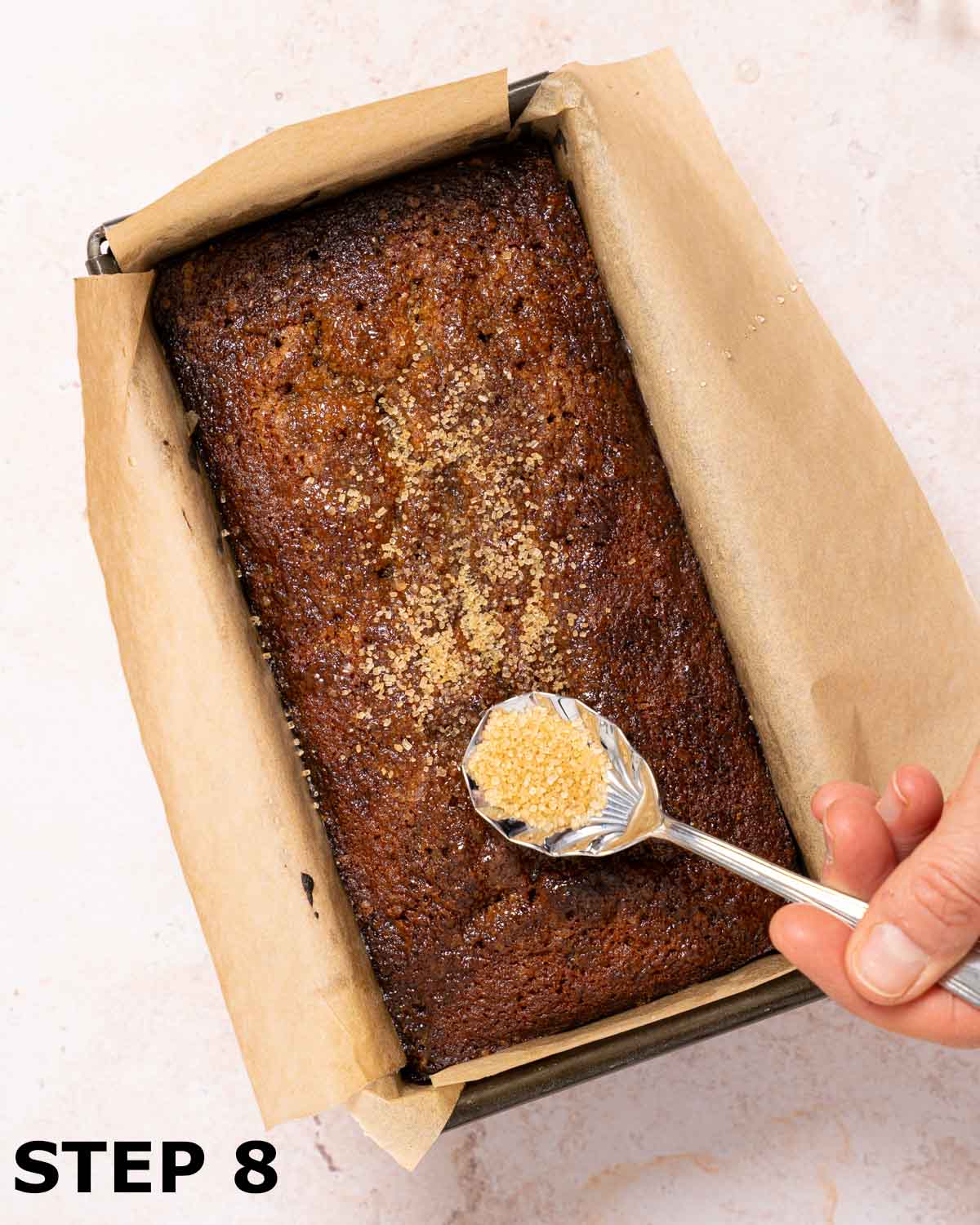 A person spooning Demerara sugar over the top of a date loaf cake still in the baking tin.