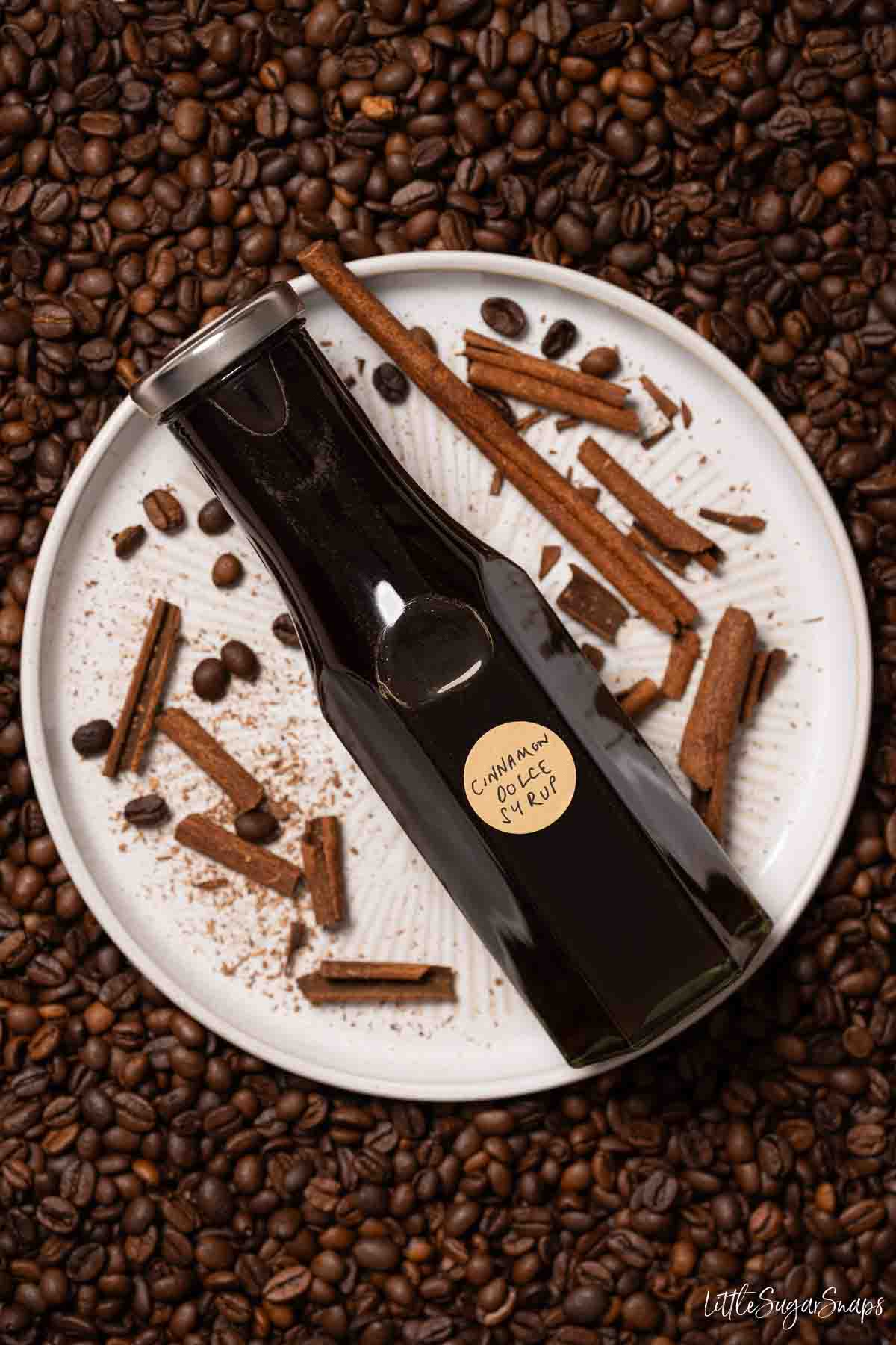 A bottle of cinnamon dolce syrup on a plate with broken cinnamon and coffee beans.