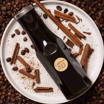 A bottle of cinnamon dolce syrup on a plate with broken cinnamon and coffee beans.
