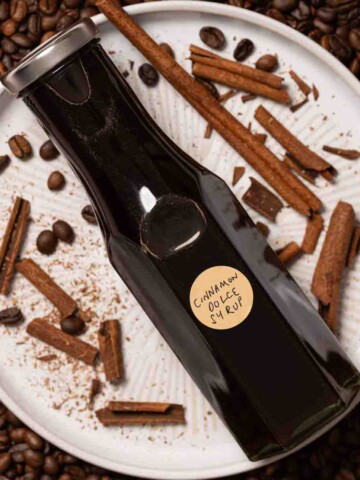 A bottle of cinnamon dolce syrup on a plate with broken cinnamon and coffee beans.