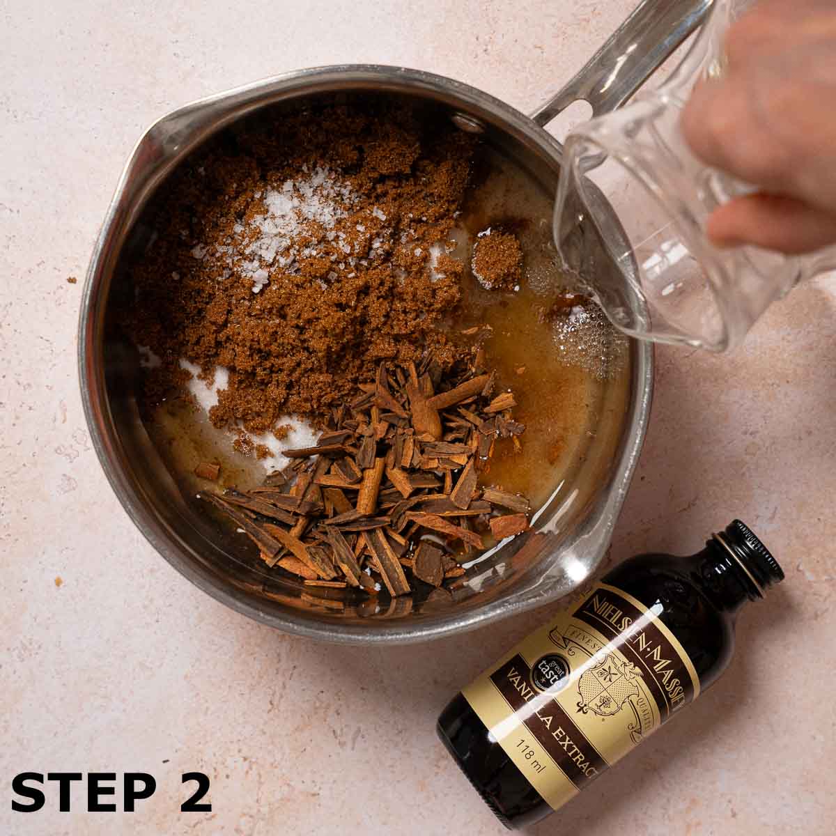 A person pouring water into a pan with sugar, crushed cinnamon and sea salt.