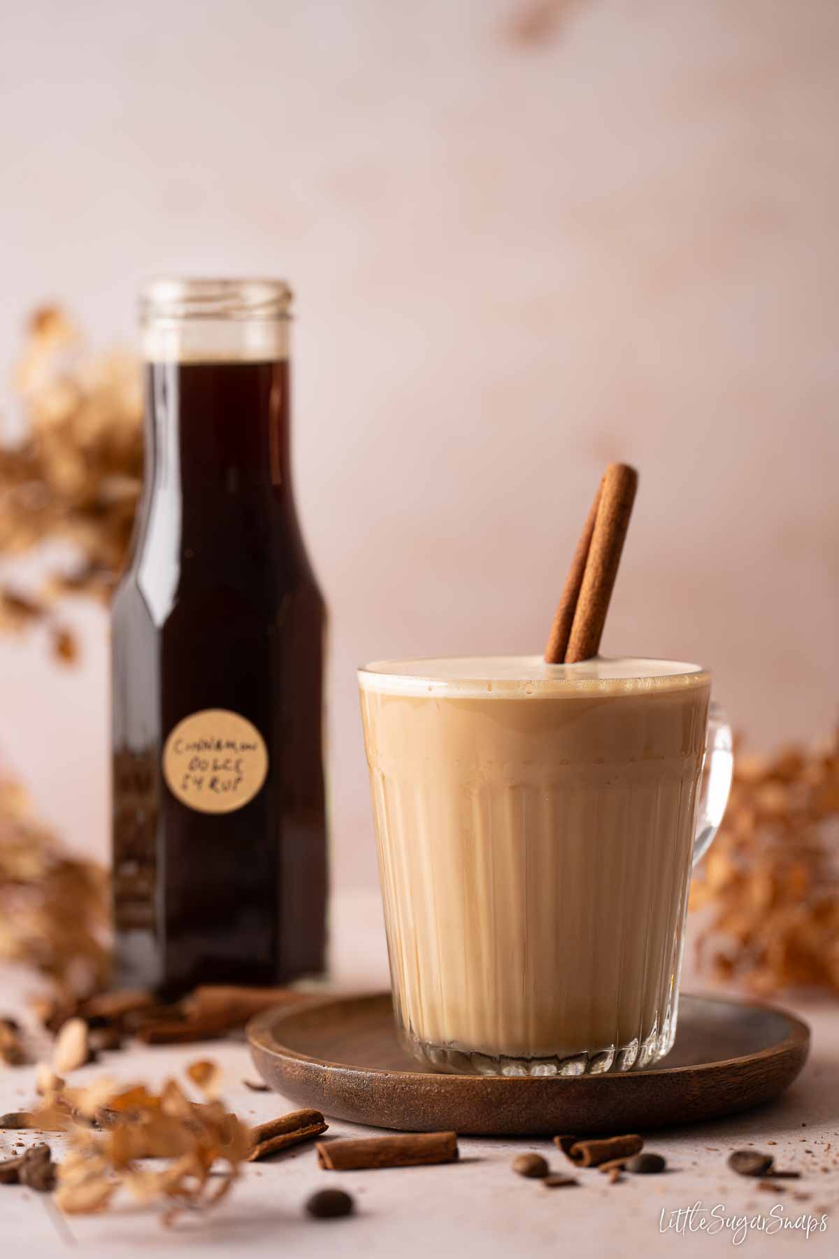 A glass mug holding cinnamon latte with cinnamon stick garnish and bottled cinnamon syrup behind it.