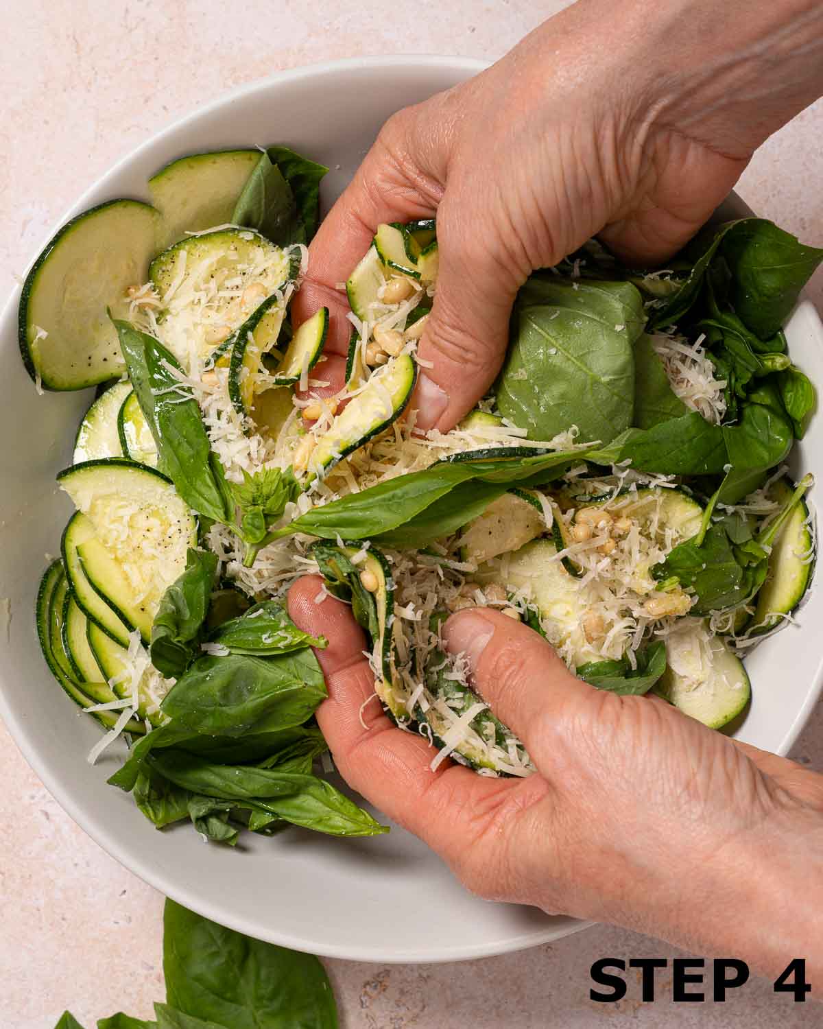 A person tossing courgette with basil leaves, grated parmesan cheese and pine nuts in a bowl.