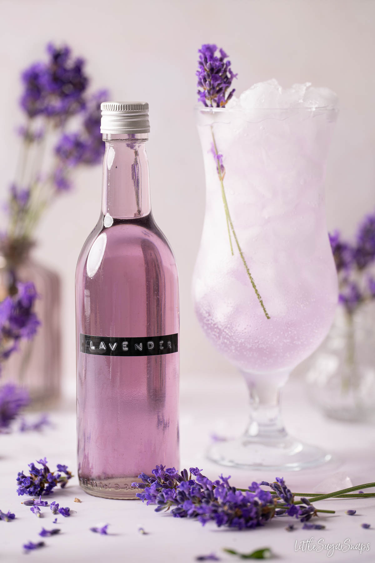A bottle of purple lavender syrup with an iced lavender drink in a hurricane glass behind it.