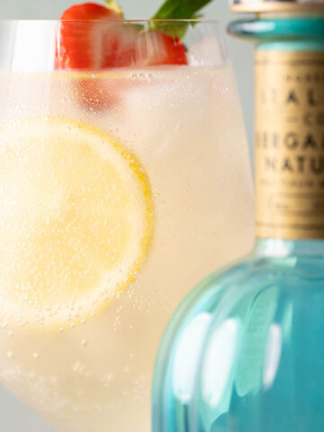 An Italicus spritz cocktail with lemon and strawberry garnish behind a bottle of Italicus liqueur.
