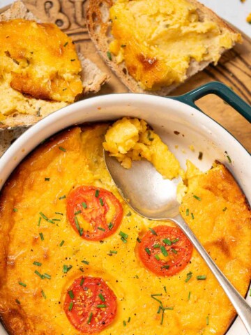 Part-served baked cheese pudding topped with sliced tomato and chives with toast on the side.