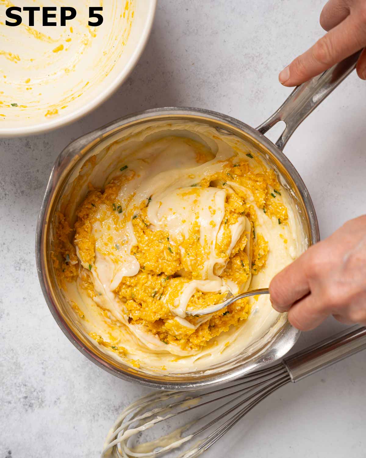 A person mixing ingredients for cheese pudding into white sauce.